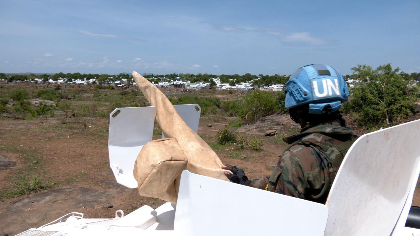 A peacekeeper peering out of the top of a tank across a grassy field. The peacekeeper is facing away from the camera.