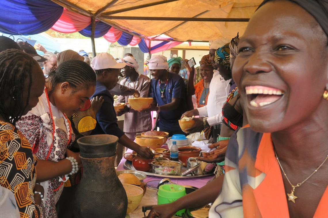 There was no lack of exciting food and cultural performances when South Sudan's official celebration of International Women's Day took place in Torit. Photos: Moses Yakudu/UNMISS A woman is smiling as she sits in front of a table full of food. There are many other women sitting at the long table which is covered with a tent.