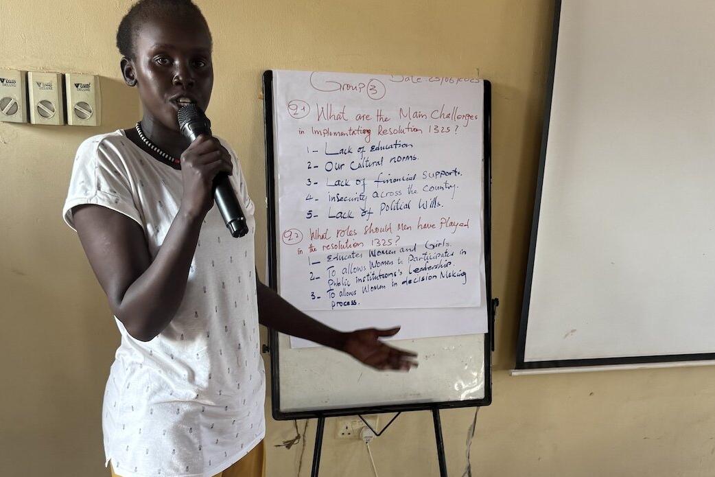 Women in Unity State have spoken out about their persistent attempts to increase and make their participation in matters related to peace and security more effective and meaningful. Person standing indoors holding a microphone and pointing toward a flip chart with handwritten notes about challenges in implementing Resolution 1325 and roles played. A blank whiteboard is positioned beside the flip chart.