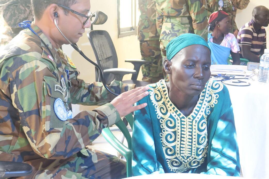 A person in camouflage uniform uses a stethoscope to examine another person wearing a turquoise garment with intricate embroidery during a medical check-up indoors