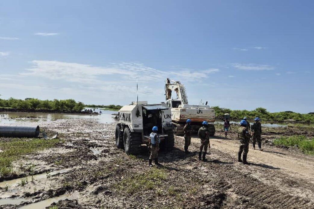 UN peacekeepers in blue helmets stand on a muddy road near heavy UN machinery and pipes, with water and greenery in the background.