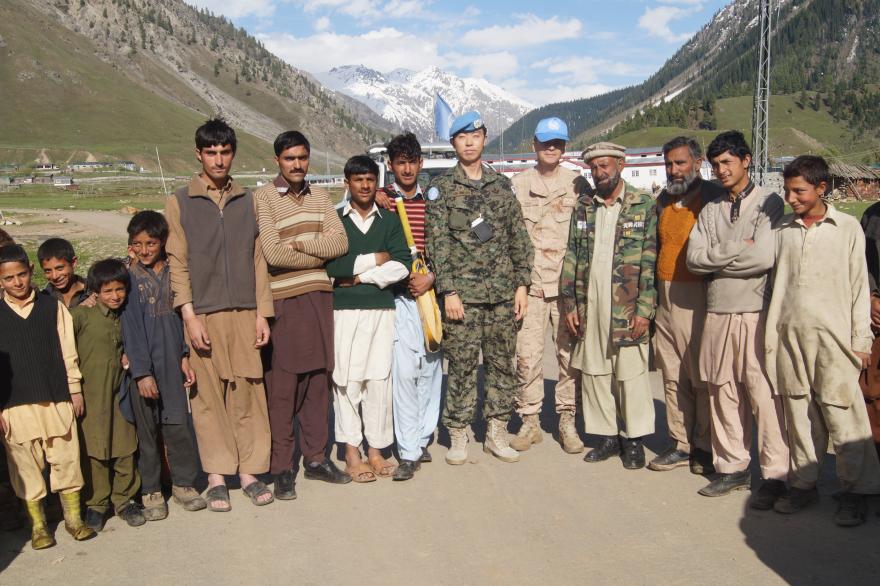 Two UN peacekeepers in blue berets stand with a group of local men and children outdoors in a mountainous area with snow-capped peaks in the background.